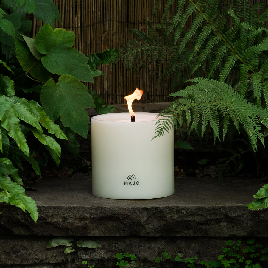 MAJO 20 cm large candle for outside in the garden. MAJO Candle lit with large flame on a step with ferns and foliage. 